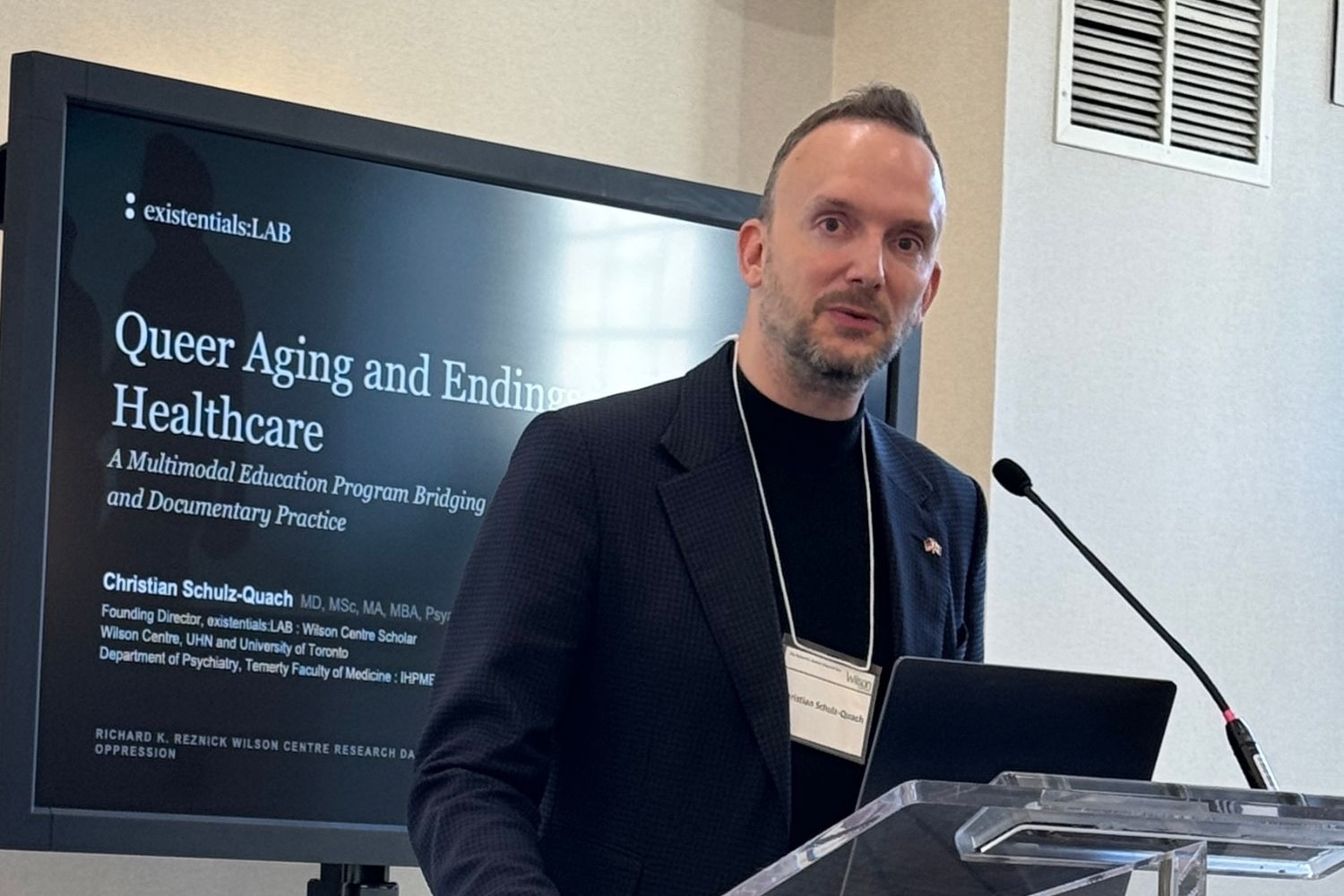 Christian Schulz-Quach speaking at a lectern beside a screen showing the title slide, Queer Aging and Endings in Healthcare.